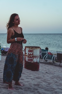 A person stands on a sandy beach near the shoreline. They are wearing colorful patterned pants and a black tank top. A rusted barrel with the word 'BEER' is visible nearby, along with some beach chairs. The ocean is calm in the background under a clear sky.