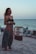 A person stands on a sandy beach near the shoreline. They are wearing colorful patterned pants and a black tank top. A rusted barrel with the word 'BEER' is visible nearby, along with some beach chairs. The ocean is calm in the background under a clear sky.