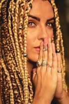A close-up of a person with long, intricately braided hair adorned with metallic beads. The person's hands are clasped together near their face, displaying multiple rings with intricate designs. The expression is contemplative and focused.