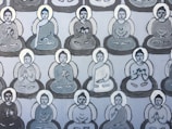 Multiple stylized figures seated in meditative postures are featured in a repeating pattern across the canvas. Each figure is encircled by a subtle halo and depicted in shades of gray and white, suggesting a calm and serene atmosphere. The figures exhibit a variety of hand gestures, commonly associated with meditation or spiritual practice.