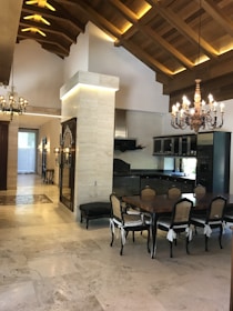 A spacious, elegantly designed dining area featuring a large wooden table surrounded by six chairs with cushioned seats. The room has a high ceiling with wooden beams and intricate chandeliers that add a touch of luxury. The floor is made of polished stone tiles, and a dark kitchen with modern appliances is visible in the background.