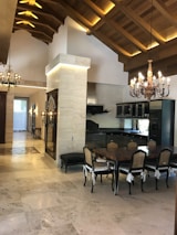 A spacious, elegantly designed dining area featuring a large wooden table surrounded by six chairs with cushioned seats. The room has a high ceiling with wooden beams and intricate chandeliers that add a touch of luxury. The floor is made of polished stone tiles, and a dark kitchen with modern appliances is visible in the background.
