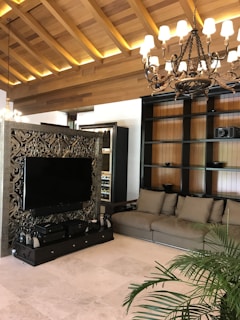 A cozy living room features a large, ornate television stand with intricate carvings, supporting a flat-screen TV. The room includes a comfortable gray sofa and a wooden bookshelf with minimal decor. Ambient lighting is provided by an elegant chandelier and warm wooden ceiling accents. A lush potted plant adds a touch of greenery to the space.