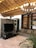A cozy living room features a large, ornate television stand with intricate carvings, supporting a flat-screen TV. The room includes a comfortable gray sofa and a wooden bookshelf with minimal decor. Ambient lighting is provided by an elegant chandelier and warm wooden ceiling accents. A lush potted plant adds a touch of greenery to the space.