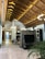 The image displays a luxurious interior with a high wooden vaulted ceiling featuring modern lighting. There is an ornate partition with a large flat-screen TV, surrounded by stylish decor. The room includes elegant chandeliers, a dining table with chairs, and plant decor, creating a sophisticated and cozy atmosphere.