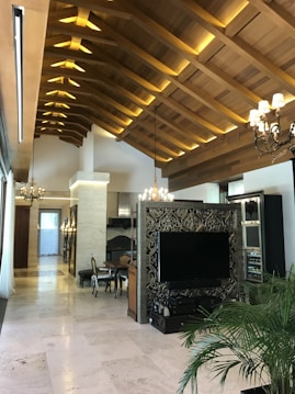 The image displays a luxurious interior with a high wooden vaulted ceiling featuring modern lighting. There is an ornate partition with a large flat-screen TV, surrounded by stylish decor. The room includes elegant chandeliers, a dining table with chairs, and plant decor, creating a sophisticated and cozy atmosphere.
