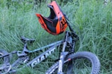 A mountain bike lying in tall green grass with a bright orange and black helmet resting on the handlebars. The bike is mostly black with white and gray accents, and it has rugged tires suitable for off-road terrain. The scene suggests a pause in an outdoor adventure or ride.