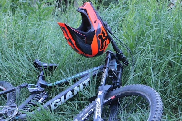 A mountain bike lying in tall green grass with a bright orange and black helmet resting on the handlebars. The bike is mostly black with white and gray accents, and it has rugged tires suitable for off-road terrain. The scene suggests a pause in an outdoor adventure or ride.