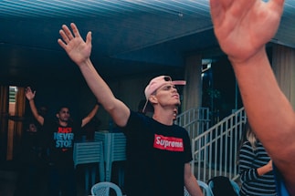 A group of people are inside a building, with a man at the center wearing a pink cap backward and a black shirt with a prominent red and white logo. He raises his right hand as if participating in an activity or event. Behind him, another person is visible with arms raised, wearing a shirt with motivational text. In the background, plastic chairs are stacked beside a staircase, and the area is dimly lit.
