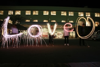 The image captures a nighttime scene with people holding sparklers that spell out the word 'Love' in the foreground. The setting is in front of a building with illuminated windows, creating a warm and inviting backdrop.