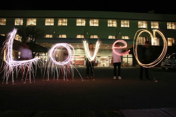 The image captures a nighttime scene with people holding sparklers that spell out the word 'Love' in the foreground. The setting is in front of a building with illuminated windows, creating a warm and inviting backdrop.