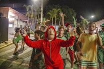 A group of people is gathered outdoors at night in a lively setting. The central figure is wearing a distinctive red costume and mask, capturing attention with a raised glass and phone in hand. Others around them are dressed casually, with many smiling and appearing to celebrate or enjoy the moment. The background includes palm trees, a building with lights, and a paved street.