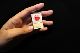 A hand holds a small rectangular pin with an image of a red lips-shaped lollipop. The background is a dark, solid color which highlights the pin and the hand. The pin has the word 'Lolita' written at the bottom.