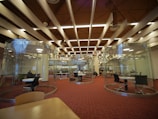 A modern library interior featuring circular glass rooms with tables and chairs inside for study or collaboration. The ceiling has distinct architectural elements with beams and lights. There are people seated at tables working or studying, and bookshelves are visible in the background.