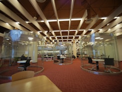 A modern library interior featuring circular glass rooms with tables and chairs inside for study or collaboration. The ceiling has distinct architectural elements with beams and lights. There are people seated at tables working or studying, and bookshelves are visible in the background.