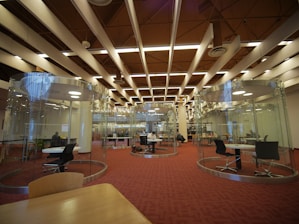 A modern library interior featuring circular glass rooms with tables and chairs inside for study or collaboration. The ceiling has distinct architectural elements with beams and lights. There are people seated at tables working or studying, and bookshelves are visible in the background.