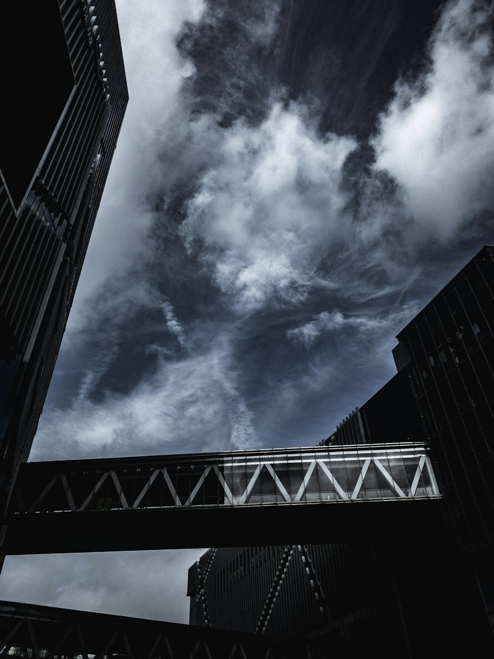 Photographie en niveaux de gris d’un pont entre deux bâtiments sous un ciel nuageux