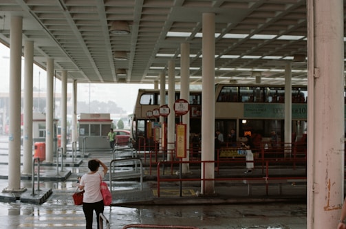A bus terminal with a double-decker bus parked next to several pillars. People are seen walking around the platform, and a woman carrying a red bag is in the foreground. The atmosphere appears wet, possibly due to rain, as evidenced by the wet pavement.