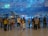 A large group of people stands in front of a massive indoor aquarium, observing aquatic life. Various types of fish swim in the blue-lit tank, with rocks and coral formations visible in the background. The spectators appear to be engaged and curious as they gaze at the marine display.