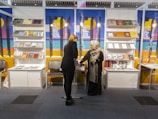 Two women are interacting at a book fair or exhibition booth. The booth displays various books and literature on shelves and tables, with colorful posters in the background. One woman is standing behind the counter, and the other is facing her, possibly inquiring about the books.