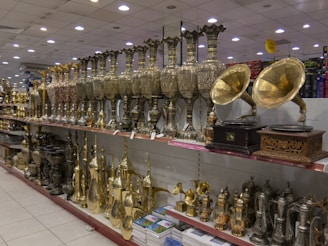 A store display featuring a variety of ornate metal objects, including large, intricately designed vases, and vintage-style gramophones with brass horns. The shelves are filled with a range of shiny and decorative items, including tea pots and other decorative vessels, reflecting a classical and traditional craftsmanship style.