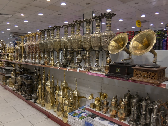 A store display featuring a variety of ornate metal objects, including large, intricately designed vases, and vintage-style gramophones with brass horns. The shelves are filled with a range of shiny and decorative items, including tea pots and other decorative vessels, reflecting a classical and traditional craftsmanship style.