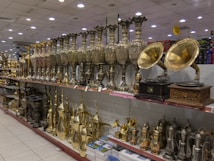 A store display featuring a variety of ornate metal objects, including large, intricately designed vases, and vintage-style gramophones with brass horns. The shelves are filled with a range of shiny and decorative items, including tea pots and other decorative vessels, reflecting a classical and traditional craftsmanship style.