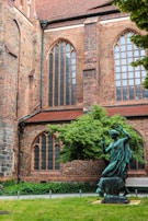 A historic brick building with tall arched windows and intricate masonry. In front, a bronze statue of a person is placed in a garden setting with a well-maintained lawn and a lush green tree.