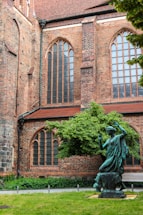 A historic brick building with tall arched windows and intricate masonry. In front, a bronze statue of a person is placed in a garden setting with a well-maintained lawn and a lush green tree.