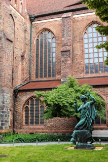 A historic brick building with tall arched windows and intricate masonry. In front, a bronze statue of a person is placed in a garden setting with a well-maintained lawn and a lush green tree.
