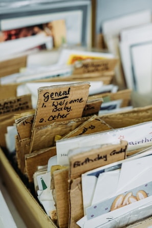 A collection of greeting cards organized into several cardboard sections in a box. Labels on the cardboard dividers indicate categories like 'General' and 'Birthday Cards.' Each section contains various cards, with some portions of the text on the cards visible.