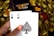 A hand holding two playing cards, an Ace of Clubs and an Ace of Spades, with a backdrop of gold Bitcoin tokens. The hand has an orange fingernail with a white Bitcoin symbol on it. The background features a stack of poker chips with a logo that reads 'Real Poker'.
