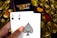 A hand holding two playing cards, an Ace of Clubs and an Ace of Spades, with a backdrop of gold Bitcoin tokens. The hand has an orange fingernail with a white Bitcoin symbol on it. The background features a stack of poker chips with a logo that reads 'Real Poker'.