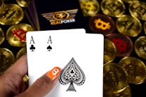 A hand holding two playing cards, an Ace of Clubs and an Ace of Spades, with a backdrop of gold Bitcoin tokens. The hand has an orange fingernail with a white Bitcoin symbol on it. The background features a stack of poker chips with a logo that reads 'Real Poker'.