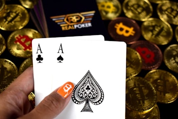 A hand holding two playing cards, an Ace of Clubs and an Ace of Spades, with a backdrop of gold Bitcoin tokens. The hand has an orange fingernail with a white Bitcoin symbol on it. The background features a stack of poker chips with a logo that reads 'Real Poker'.