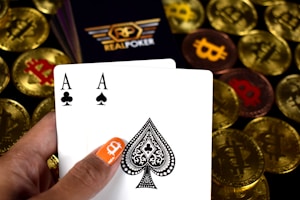 A hand holding two playing cards, an Ace of Clubs and an Ace of Spades, with a backdrop of gold Bitcoin tokens. The hand has an orange fingernail with a white Bitcoin symbol on it. The background features a stack of poker chips with a logo that reads 'Real Poker'.