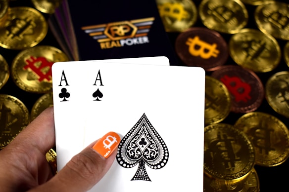 A hand holding two playing cards, an Ace of Clubs and an Ace of Spades, with a backdrop of gold Bitcoin tokens. The hand has an orange fingernail with a white Bitcoin symbol on it. The background features a stack of poker chips with a logo that reads 'Real Poker'.