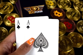 A hand holding two playing cards, an Ace of Clubs and an Ace of Spades, with a backdrop of gold Bitcoin tokens. The hand has an orange fingernail with a white Bitcoin symbol on it. The background features a stack of poker chips with a logo that reads 'Real Poker'.