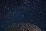 A night sky filled with countless stars, with a large radio telescope dish silhouetted against the celestial background.