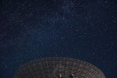 A night sky filled with countless stars, with a large radio telescope dish silhouetted against the celestial background.
