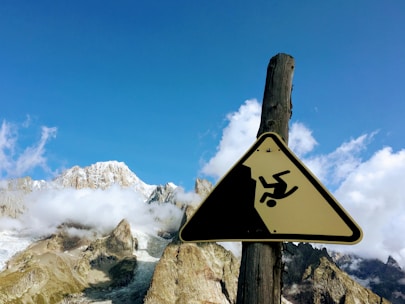 A triangular warning sign depicting a person falling off a cliff is mounted on a wooden post. Behind the sign is a breathtaking view of snow-capped mountains with patches of clouds against a clear blue sky.