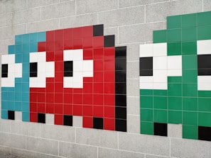 Three stylized pixelated characters resembling ghosts from the classic Pac-Man video game are depicted on a tiled wall. Each character is made from square tiles, with the left one in light blue, the center one in red, and the right one in green. The characters have large white eyes with black pupils and are set against a beige brick wall.