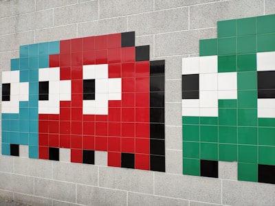 Three stylized pixelated characters resembling ghosts from the classic Pac-Man video game are depicted on a tiled wall. Each character is made from square tiles, with the left one in light blue, the center one in red, and the right one in green. The characters have large white eyes with black pupils and are set against a beige brick wall.