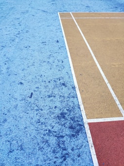 A sports court surface featuring different colored sections with textured patterns. The court includes a large blue area with subtle speckling, a mustard yellow area with crisp white lines, and a smaller red rectangle. The geometric layout forms a sharp contrast between the colors.