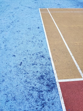 A sports court surface featuring different colored sections with textured patterns. The court includes a large blue area with subtle speckling, a mustard yellow area with crisp white lines, and a smaller red rectangle. The geometric layout forms a sharp contrast between the colors.