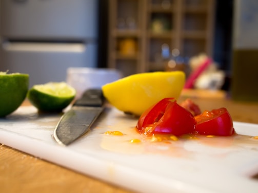 A kitchen scene featuring cut vegetables and a knife on a cutting board. Half of a lime, a yellow bell pepper, and sliced red tomatoes are visible. The background consists of a blurry kitchen shelf and other kitchen items.