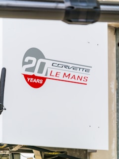 A sign featuring a design with the text '20 Years Corvette Le Mans' in bold red and gray colors. The background is white, and there is a partial view of a black object slightly obscuring part of the sign.