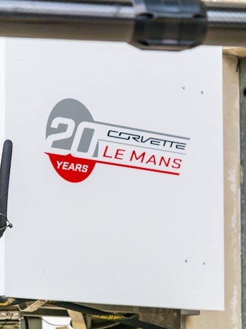 A sign featuring a design with the text '20 Years Corvette Le Mans' in bold red and gray colors. The background is white, and there is a partial view of a black object slightly obscuring part of the sign.