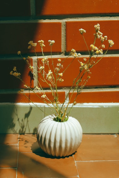 A softly lit corner featuring a minimalist ceramic vase holding delicate dried flowers on a light wooden table.