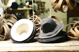 A collection of hats is displayed on a wooden surface, surrounded by loops of material. The hats are primarily dark in color, with one lighter hat showing its underside. The setting appears to be a workshop, as suggested by the blurred background with a person working and various tools visible.